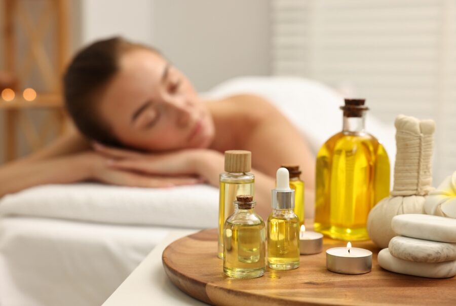 A person rests on a massage table in a spa setting, eyes closed, surrounded by an aura of tranquility. Nearby, bottles of Salt & Sage healing oils and small stones adorn a wooden tray. The soft lighting enhances the calming atmosphere, inviting deep relaxation and rejuvenation.