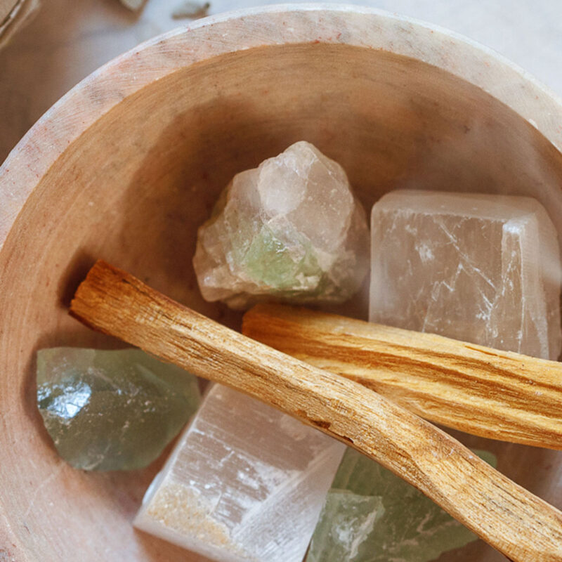 Wooden bowl filled with assorted healing crystals and palo santo sticks, placed on a light stone surface.