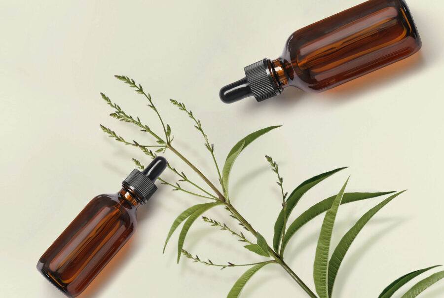 Two amber glass dropper bottles are placed diagonally on a light background alongside green leafy branches. The arrangement creates a natural and minimalist aesthetic.