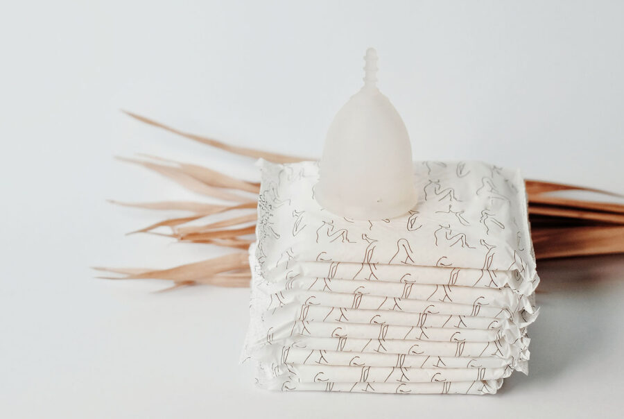 A white menstrual cup sits on top of a stack of white sanitary pads with abstract line patterns. Dried leaves are placed next to the stack, against a plain white background.