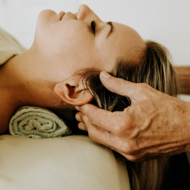 A person lies on their back with closed eyes, resting their head on a towel. Another persons hands gently massage their scalp. The setting is calm and relaxing.