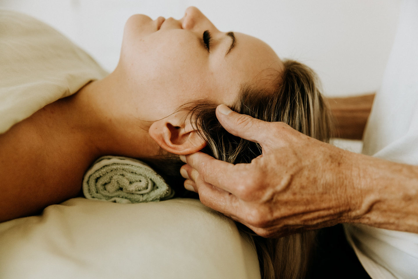 A person lies on their back with closed eyes, resting their head on a towel. Another persons hands gently massage their scalp. The setting is calm and relaxing.