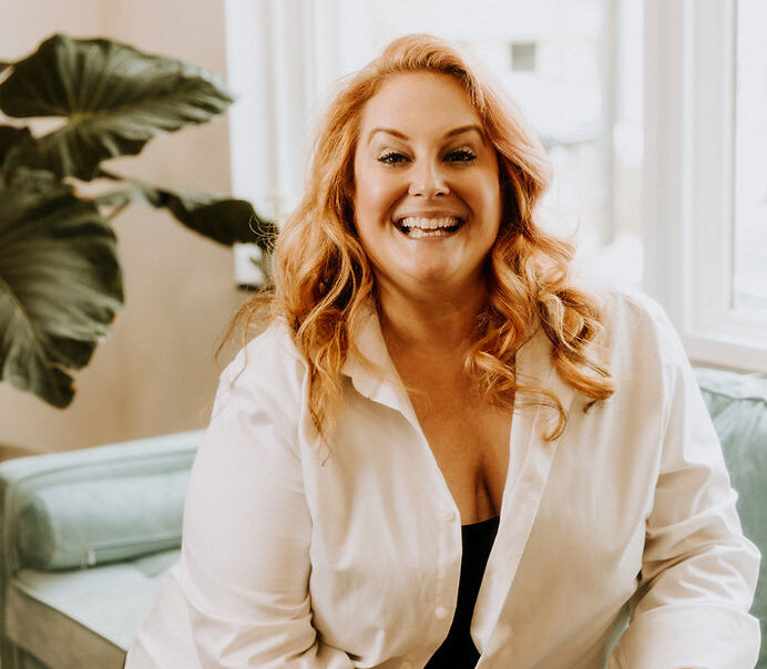 A person with long red hair, a member of the Salt & Sage Team, smiles warmly while seated on a light green couch. Wearing a white shirt in a room with large windows and a plant in the background, they exude an inviting presence.