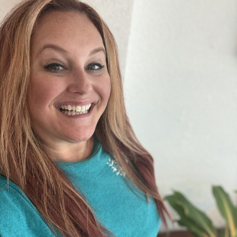 Smiling person in turquoise sweater with plant backdrop.
